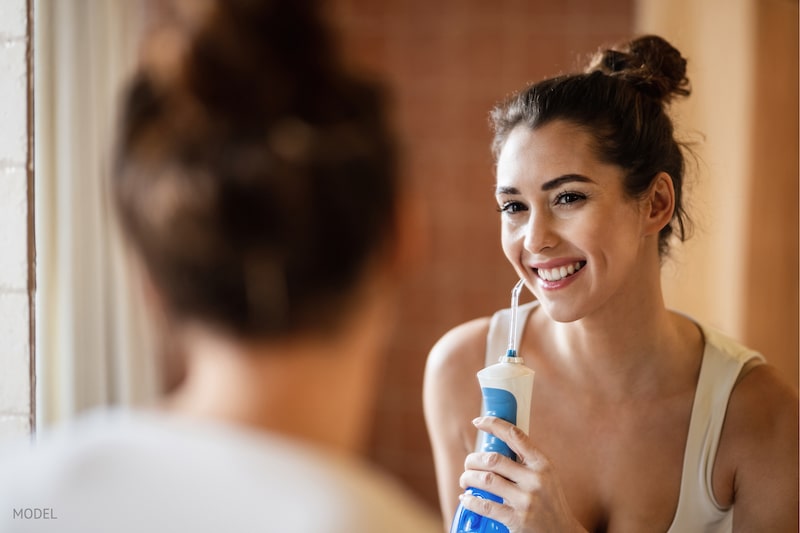 woman using dental water flosser while cleaning teeth and looking herself in bathroom mirror.