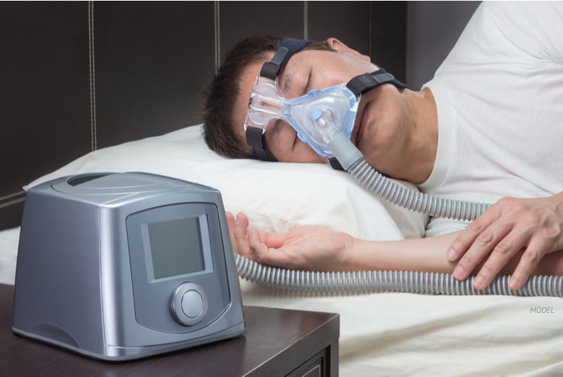Man sleeping with CPAP machine mask strapped to his face and machine in the foreground.